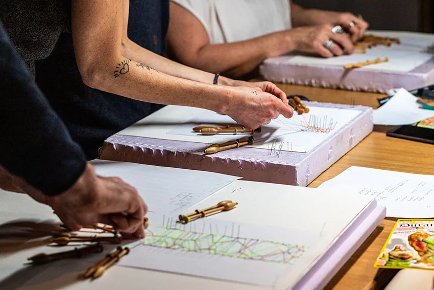 3 paires de mains manipulant les fuseaux de dentelle avec des fils colorés vert, jauen orange
